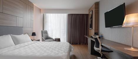 A hotel room with a large bed, a desk with a chair, a flat-screen TV, and a lamp.