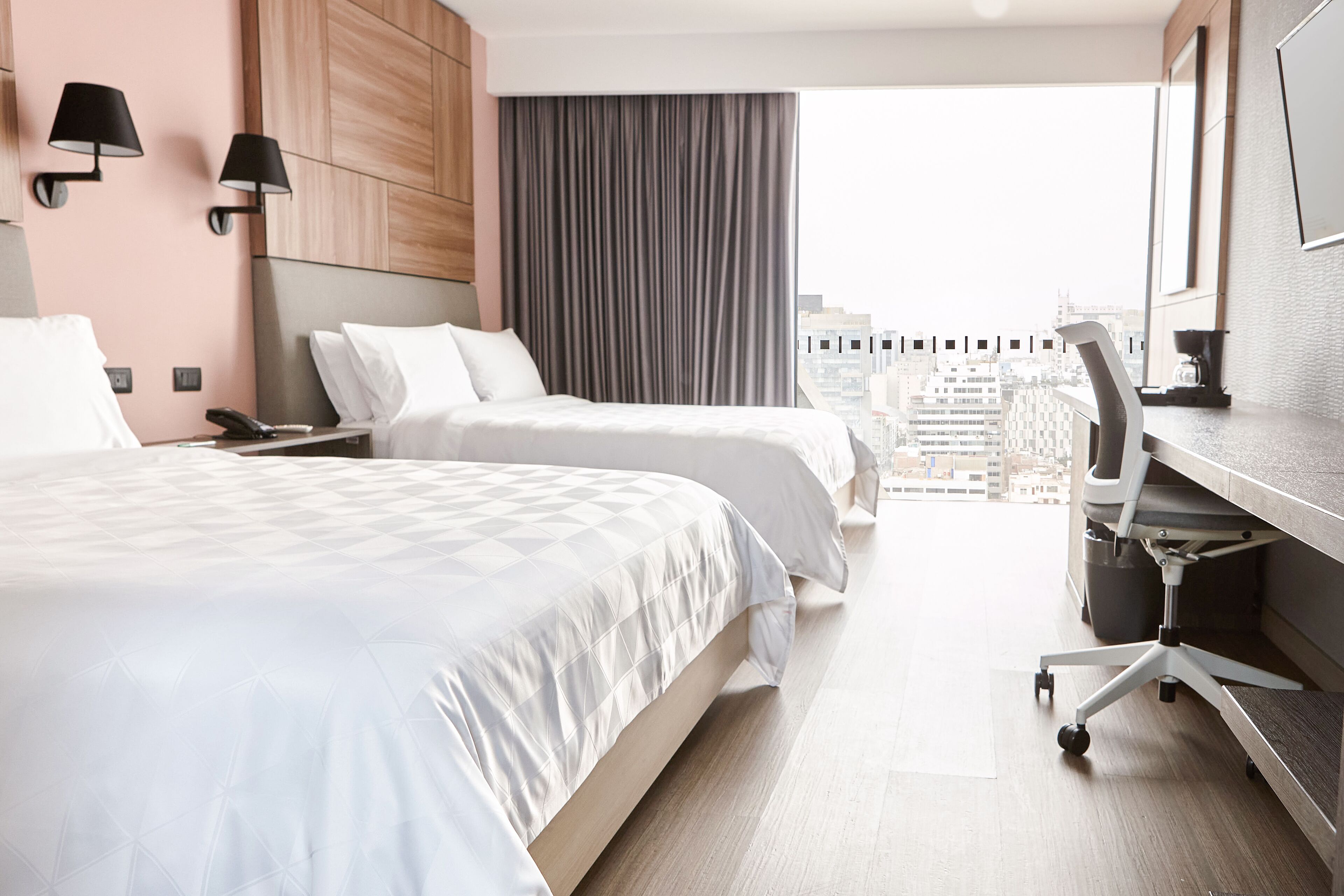 Una habitación de hotel con dos camas, un escritorio con silla, un ventanal con vistas a edificios y un paisaje urbano.