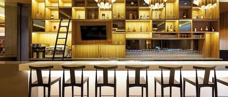 Una zona de bar con una barra larga, varios taburetes y estantes llenos de botellas y copas.