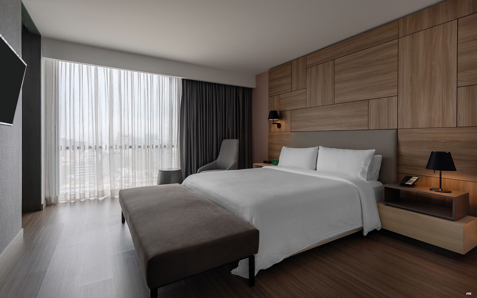 A modern hotel room with a large bed, a bench, a desk with a lamp, and a view of the city through sheer curtains.