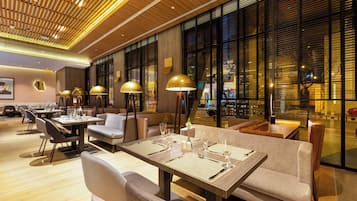 Un comedor moderno con mesas dispuestas para dos, iluminación ambiental y vistas amplias a través de grandes ventanales.