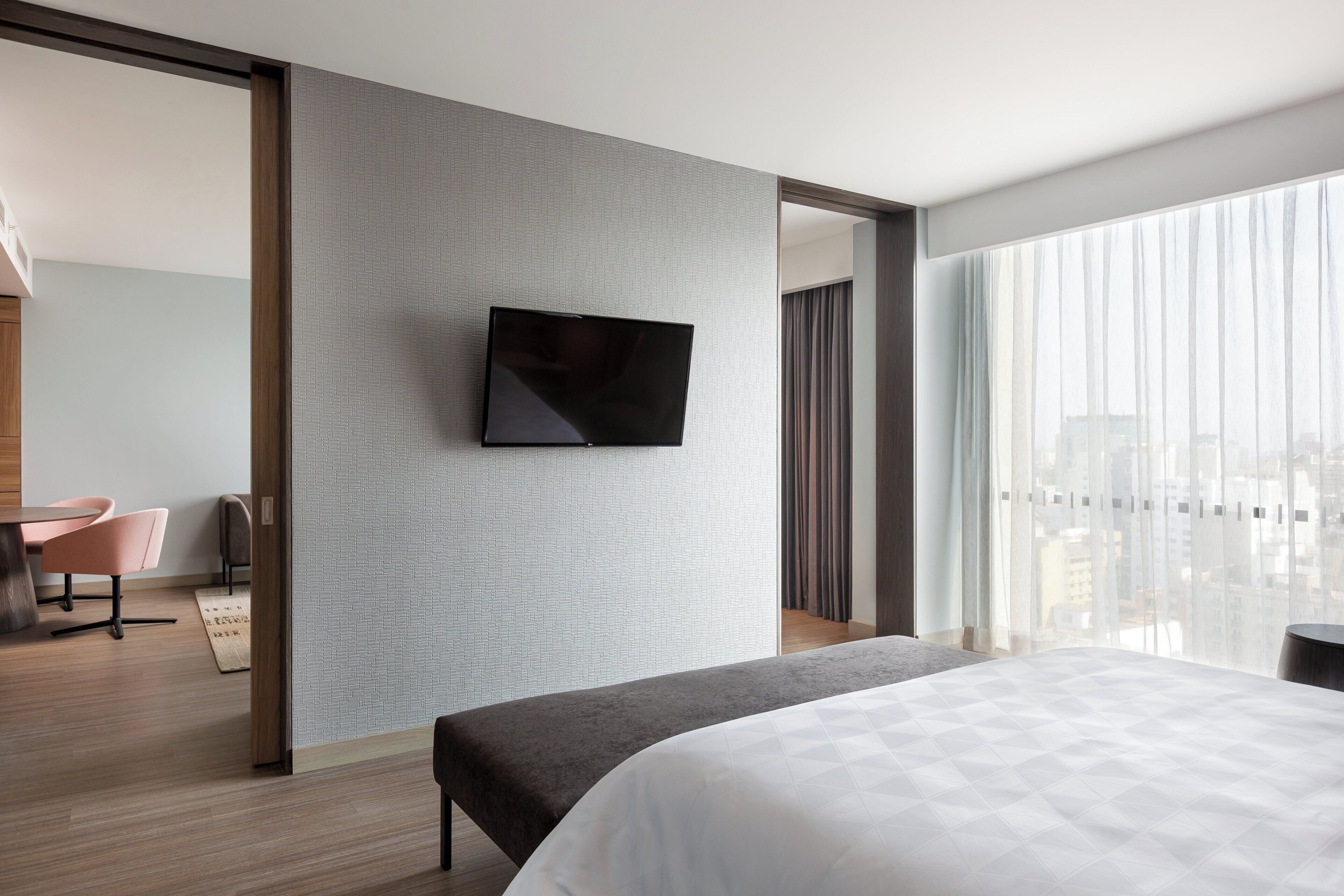 Habitación de hotel moderna con una cama grande, un televisor de pantalla plana montado en una pared con textura, una zona de estar con una silla y una mesa, y vistas de la ciudad a través de amplios ventanales.