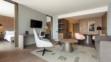 A modern hotel room with a bed, a desk, a television, and a seating area with a table and chairs.