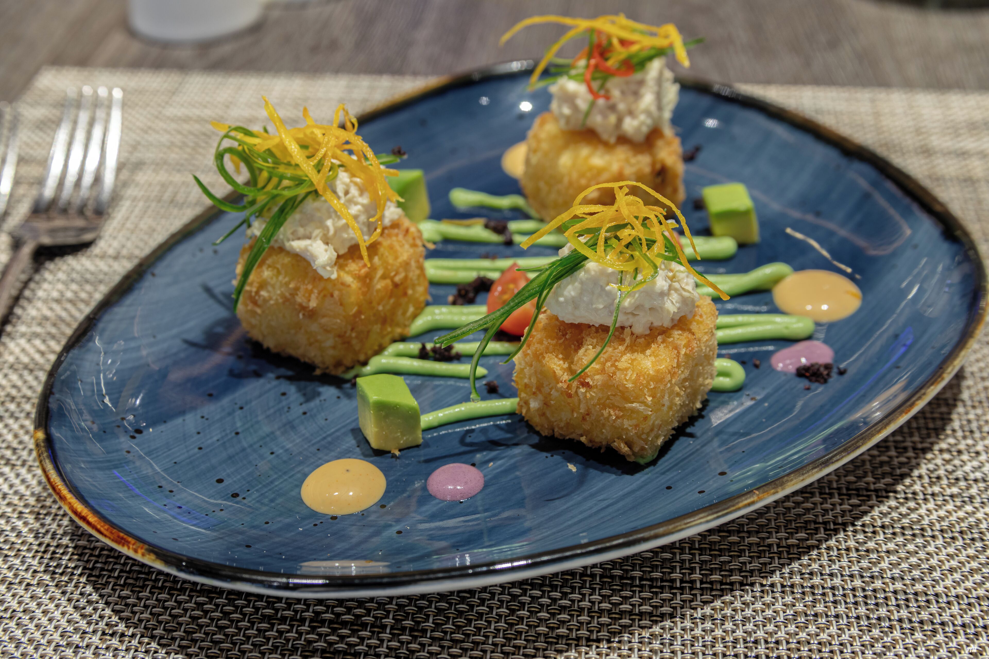 A plate of food with three spherical items topped with a white substance and garnished with green and yellow elements, accompanied by green sauce and small round items.