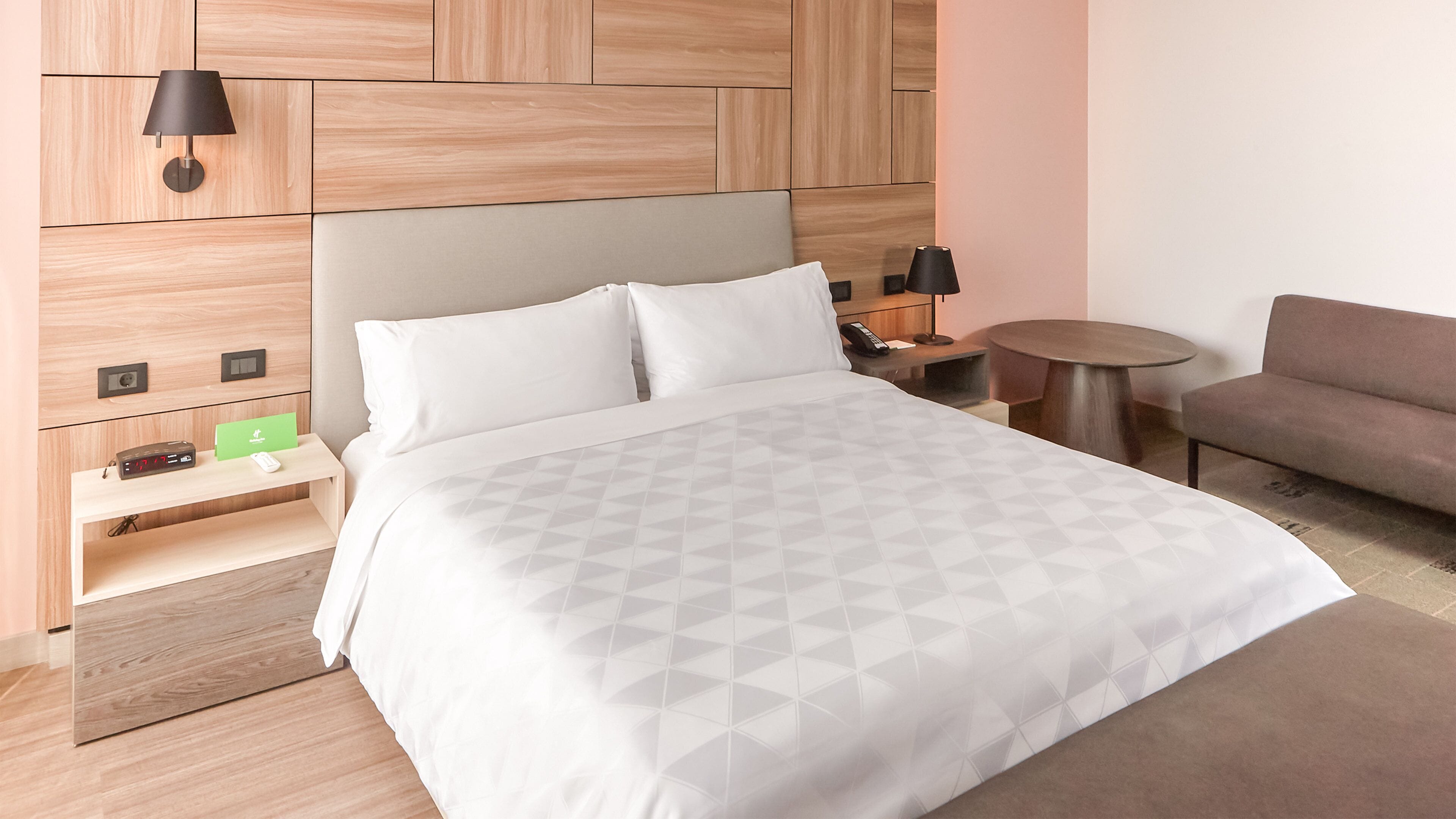 A modern hotel room with a large bed, a bedside table with a lamp, a small round table, and a sofa.