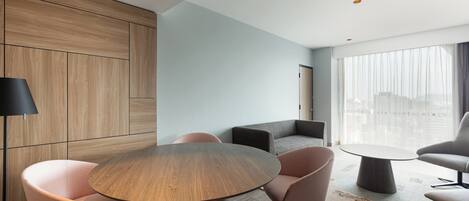 A modern hotel room with a round wooden table, cushioned chairs, a sofa, and a large window with a view.