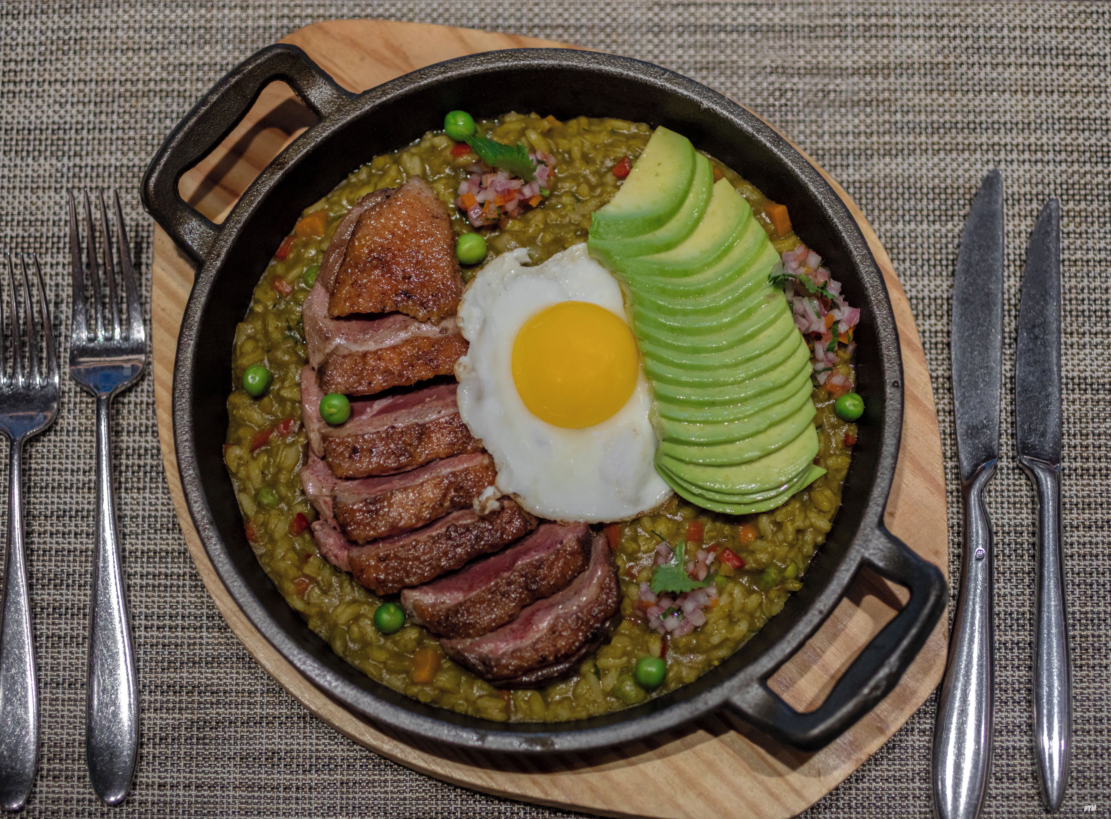 A skillet with sliced meat, an egg, and avocado on a textured tablecloth.