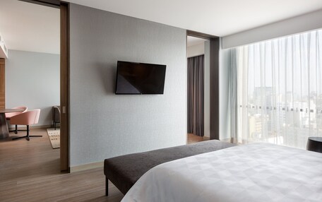 A modern hotel room with a large bed, a flat-screen TV mounted on the wall, a dining area with a table and chairs, and a view of the city through large windows.