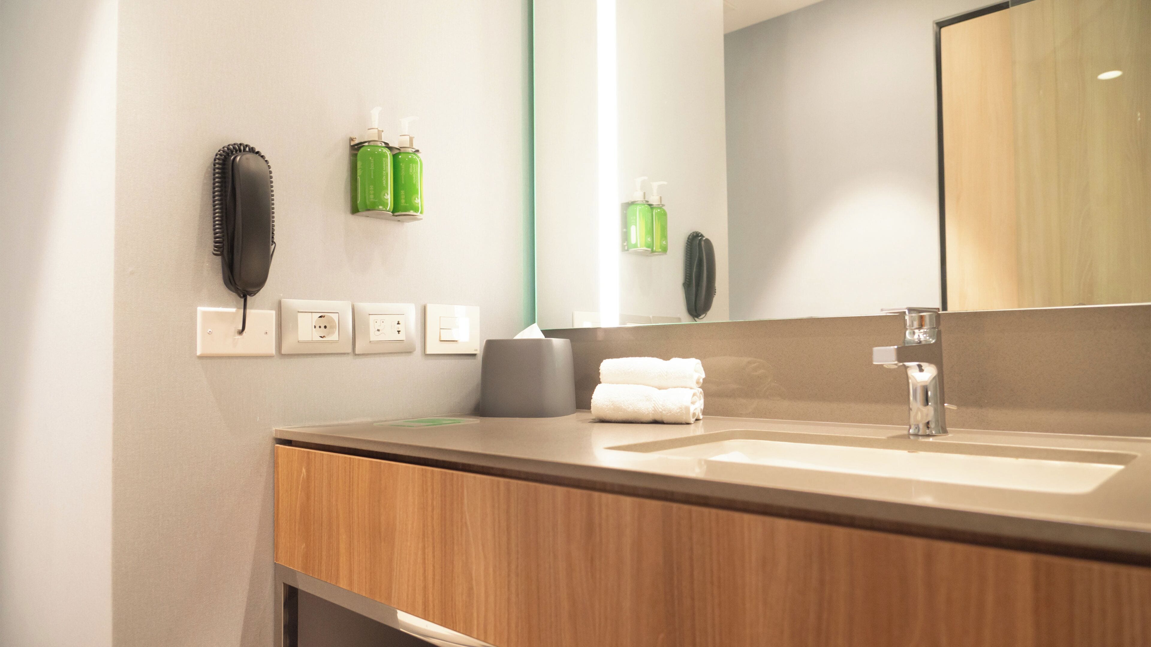 Een badkamer met een houten wastafelmeubel, een spiegel, een wasbak en twee groene flesjes handdesinfectiemiddel.