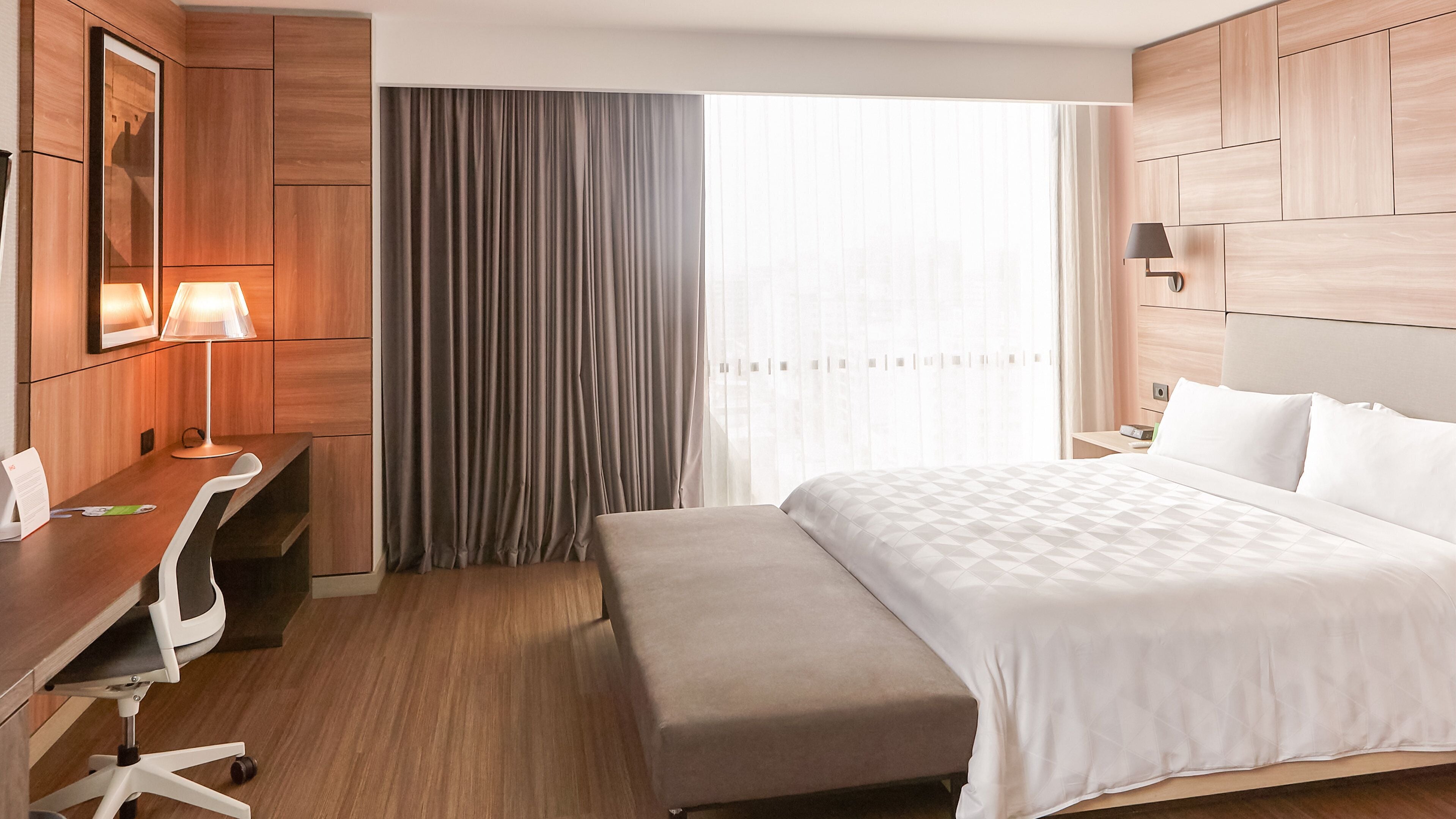 A hotel room with a bed, a desk with a lamp, a chair, a bench, and a large window with curtains.