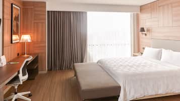 Ein Hotelzimmer mit einem Bett, einem Schreibtisch mit Lampe, einem Sessel, einer Bank und einem großen Fenster mit Vorhängen.