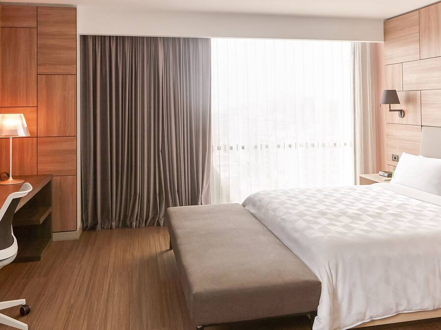 Ein Hotelzimmer mit einem Bett, einem Schreibtisch mit Lampe, einem Sessel, einer Bank und einem großen Fenster mit Vorhängen.