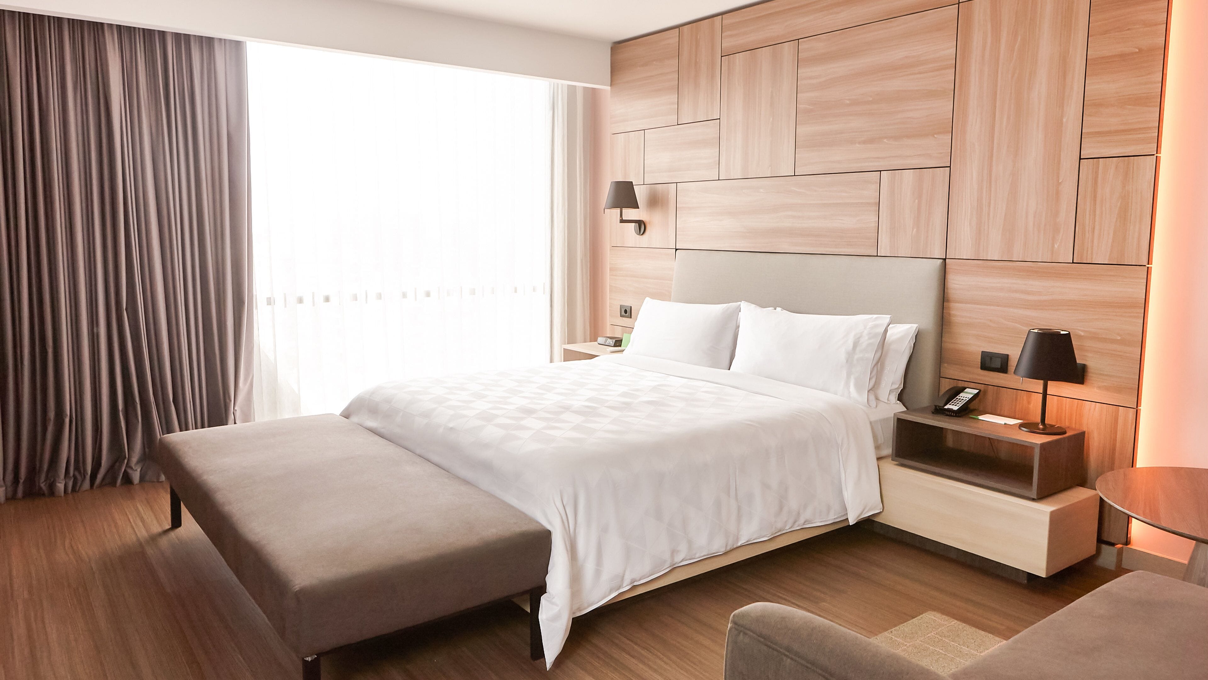 Una habitación de hotel moderna con una cama grande, una mesita de noche con lámpara, una mesita auxiliar con teléfono, un banco y un ventanal con cortinas.