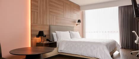 A modern hotel room with a large bed, a bedside table with a lamp, a small round table, and a large window with curtains.