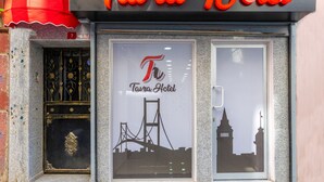 Een hotelingang met een bord waarop 'Tavra Hotel' staat en een logo met de silhouetten van een brug en een toren.