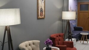 A room with a grey wall, a framed artwork, a beige armchair, a red armchair, a small round table with a vase, and a floor lamp. - Tavra Hotel (Istanbul)