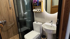 Een moderne badkamer met een douchecabine van glas, een toilet, een wastafel en een houten plank met toiletartikelen.