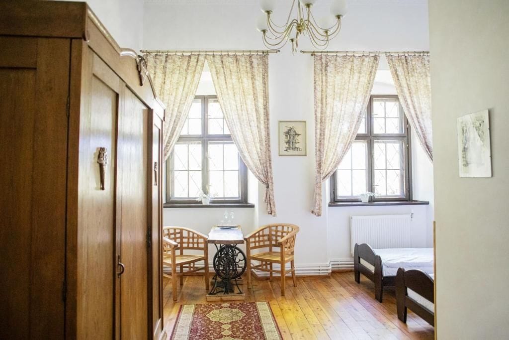 Una habitación con un armario de madera, dos ventanas con cortinas, una mesa con sillas, un radiador y una cama.