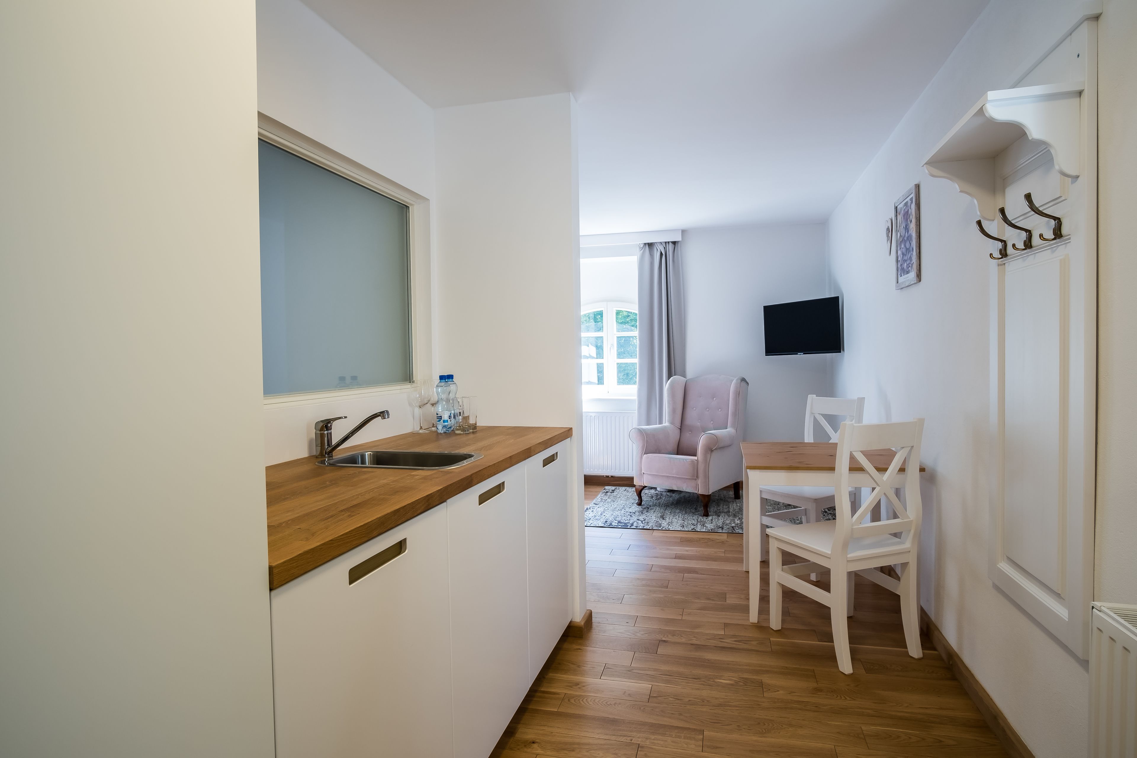 A compact kitchen with a wooden countertop, a sink, and a window. There is a dining area with a table and chairs, and a television mounted on the wall.