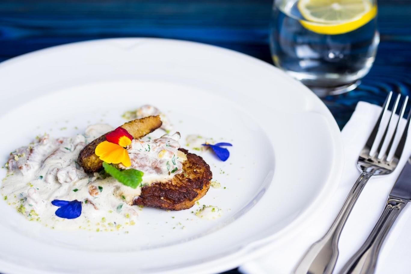 Un plato blanco con un trozo de carne, una cucharada de salsa y una guarnición de flores y hierbas.