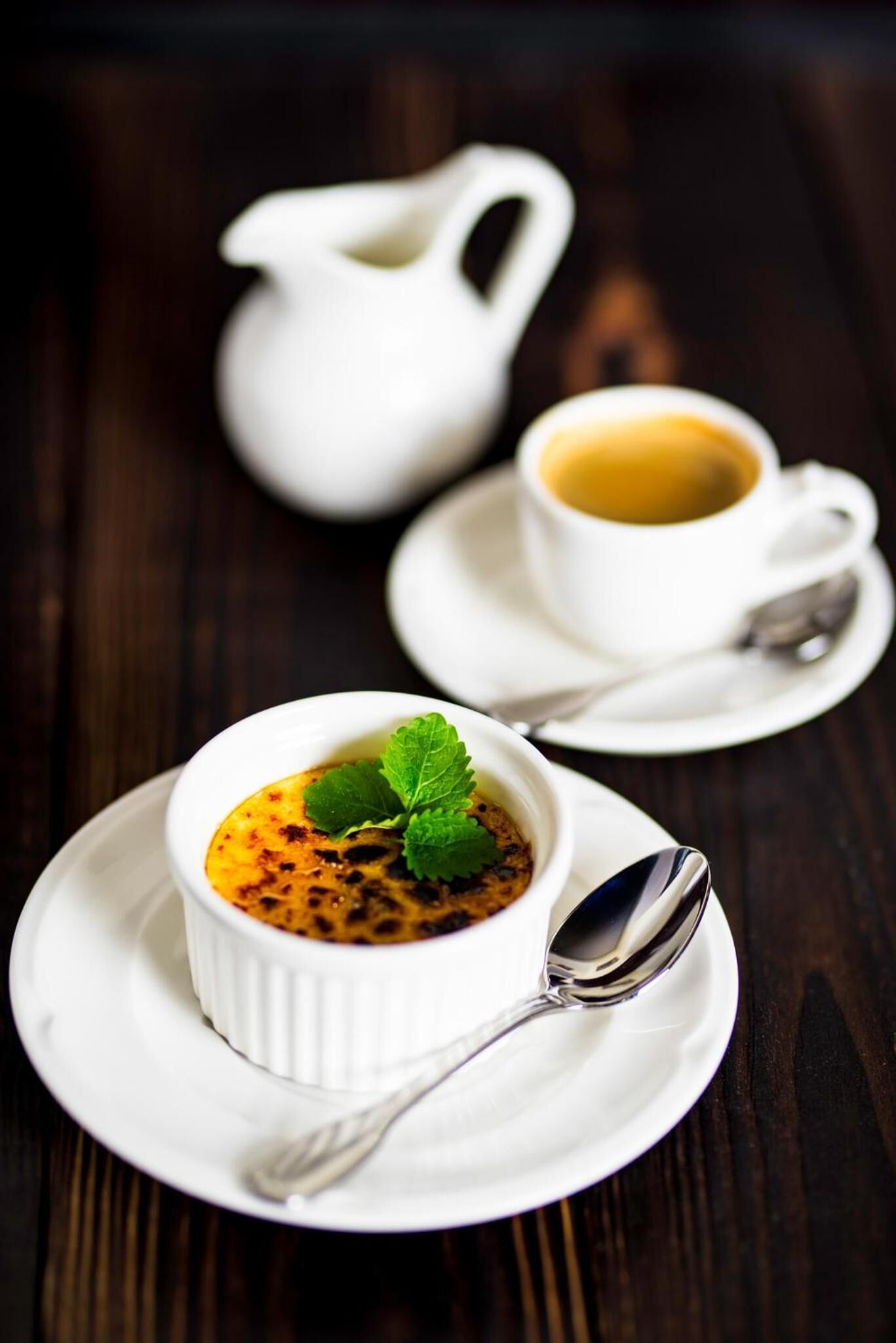 Un postre en un recipiente blanco sobre un plato blanco, junto con una cuchara, una jarra blanca y una taza de café en un platito.