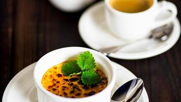 Un postre en un recipiente blanco sobre un plato blanco, junto con una cuchara, una jarra blanca y una taza de café en un platito.