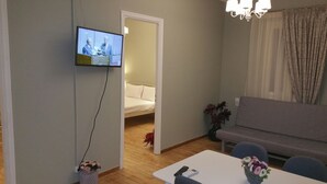 A living room with a TV mounted on the wall, a sofa, a dining table with a vase of flowers, and a bed in the adjacent room. - Feanis House (Lefkada)