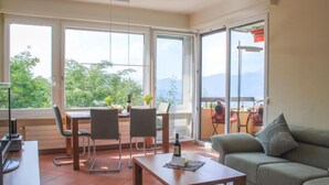 A modern living room with a dining area, large windows, and a view of greenery and mountains. - Residenza Monte Bré - Apt.004 Haus1 by Interhome (Aldesago)