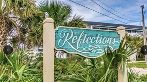 A sign with the word "Reflections" surrounded by lush greenery and a building in the background. - Oceanfront condo w/direct beach access & 180 degree views from your private deck (Garden City Beach)