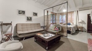 Een woonkamer met een bank, fauteuil en salontafel. Er staat een bed met raam en lamp.