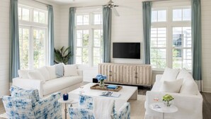 Ein geräumiges Wohnzimmer mit einem weißen Sofa, blauen, gemusterten Sesseln, einem Couchtisch und einem Fernseher auf einer Holzeinrichtung.