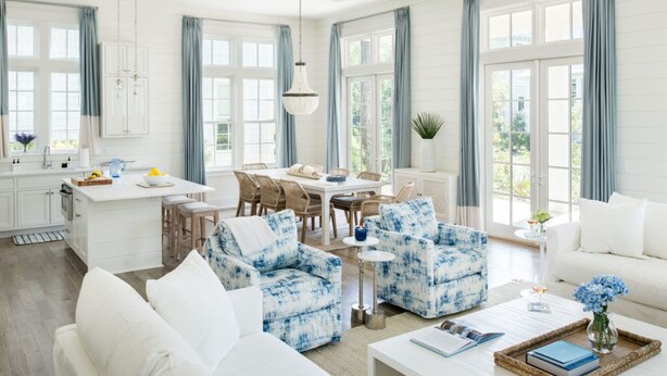 Ein geräumiges Wohnzimmer mit einem weißen Sofa, blauen, gemusterten Sesseln, einem Essbereich mit einem weißen Tisch und Stühlen sowie großen Fenstern mit blauen Vorhängen.