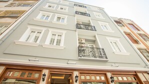 Un edificio de varios pisos con fachada gris claro, detalles blancos y un balcón con barandillas negras ornamentadas.