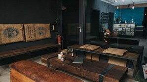 Un espacio lounge moderno y acogedor con bancos y mesas de madera, un extintor y vistas al área del bar a través de un panel de vidrio.