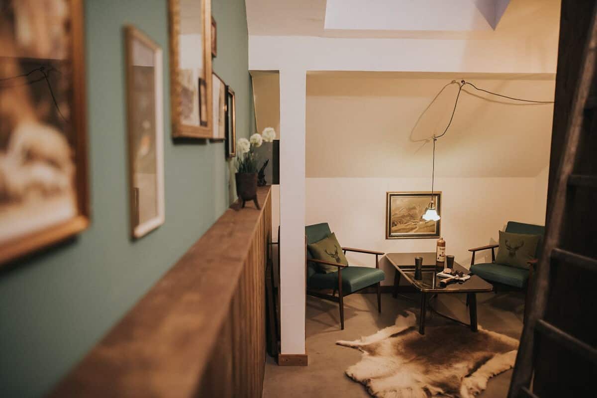 Una habitación acogedora con un banco de madera, dos sillas verdes, una mesita con una lámpara y un manto de piel.
