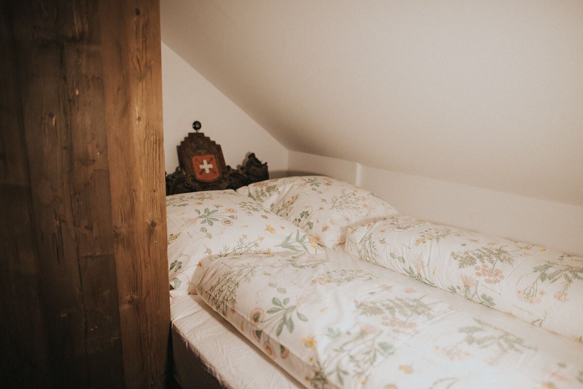 A small, cozy room with a wooden door, a bed with floral bedding, and a small wooden chest with a red cross emblem.