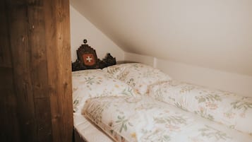 A small, cozy room with a wooden door, a bed with floral bedding, and a small wooden chest with a red cross emblem.