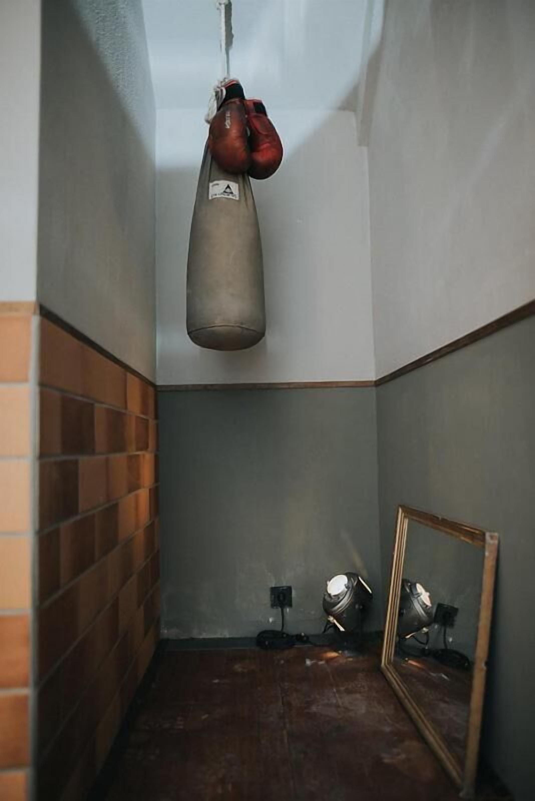 Una habitación estrecha con un saco de boxeo, un espejo y una pequeña mesa con una silla.