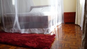 A bedroom with a bed under a mosquito net, a red rug, wooden flooring, and a small wooden cabinet. - Sheema Villas (Nairobi)