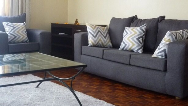 A living room with a grey sofa, patterned cushions, a glass coffee table, and a white rug. - Sheema Villas (Nairobi)