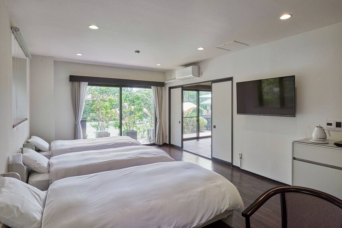 Une chambre d’hôtel avec trois lits, une télévision, un bureau avec une bouilloire et une vue sur la verdure environnante.