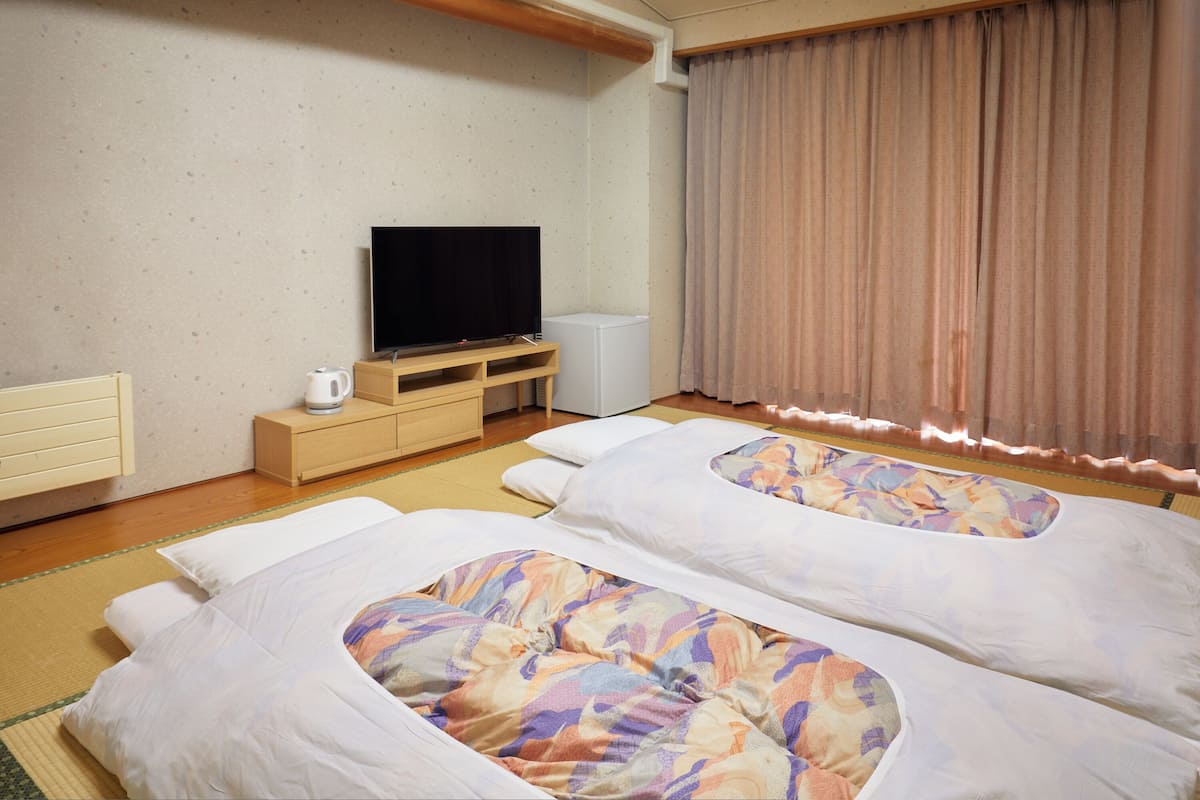 Habitación de hotel con dos camas, televisor, un mueble blanco pequeño y un ventanal con cortinas.