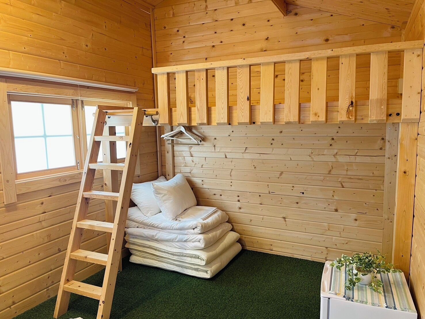 A wooden cabin room with a ladder, a stack of white bedding, a small plant, and a window.