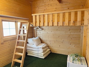 Interior de una cabaña de madera con una escalera, ropa de cama blanca apilada, una planta pequeña y una ventana.