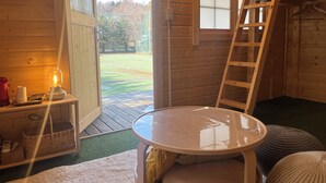 Interior de una cabaña de madera con una mesa, una escalera, un ventanal y vistas a un campo cubierto de hierba.