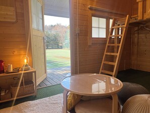 Interior de uma cabana de madeira com uma mesa, uma escada, uma janela e uma porta que dá para um gramado.
