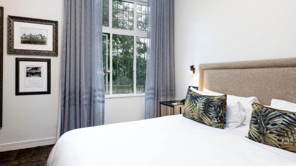 Ein Hotelzimmer mit einem großen Bett, einem Nachttisch, einem Fenster mit Vorhängen und gerahmten Bildern an der Wand.