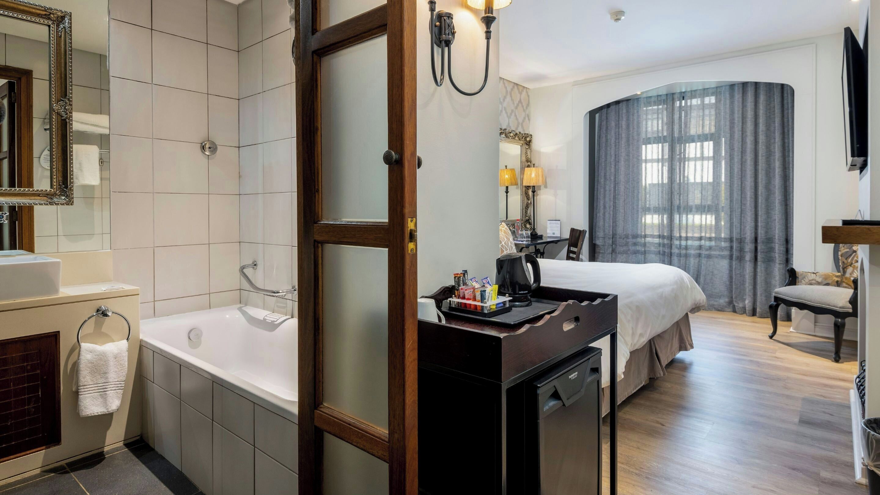 A hotel room with a bathtub, a desk with a coffee maker, a bed, and a large window with curtains.