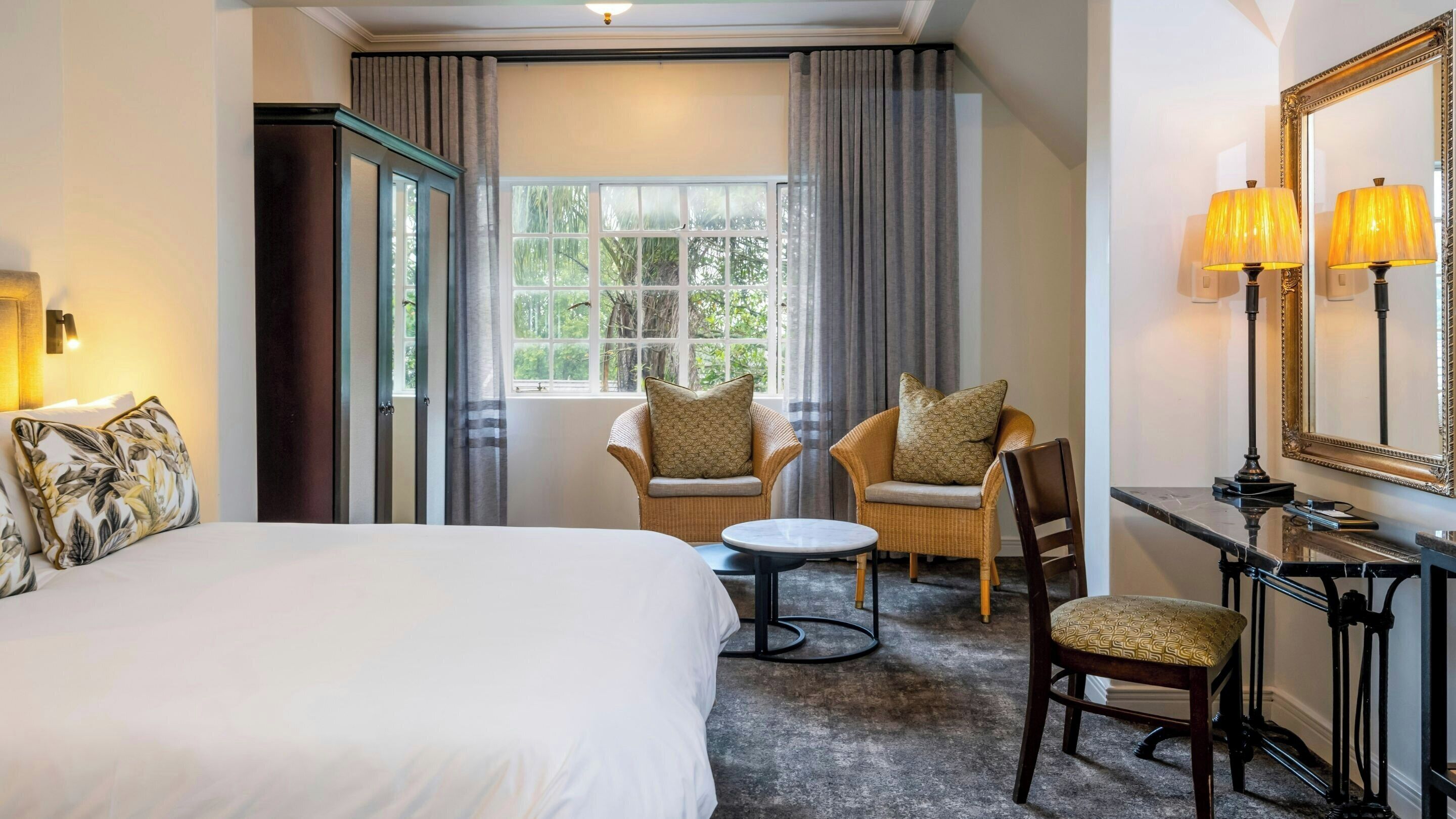 Habitación de hotel con cama, dos sillas, una mesita, escritorio con lámpara, espejo y ventana con cortinas.