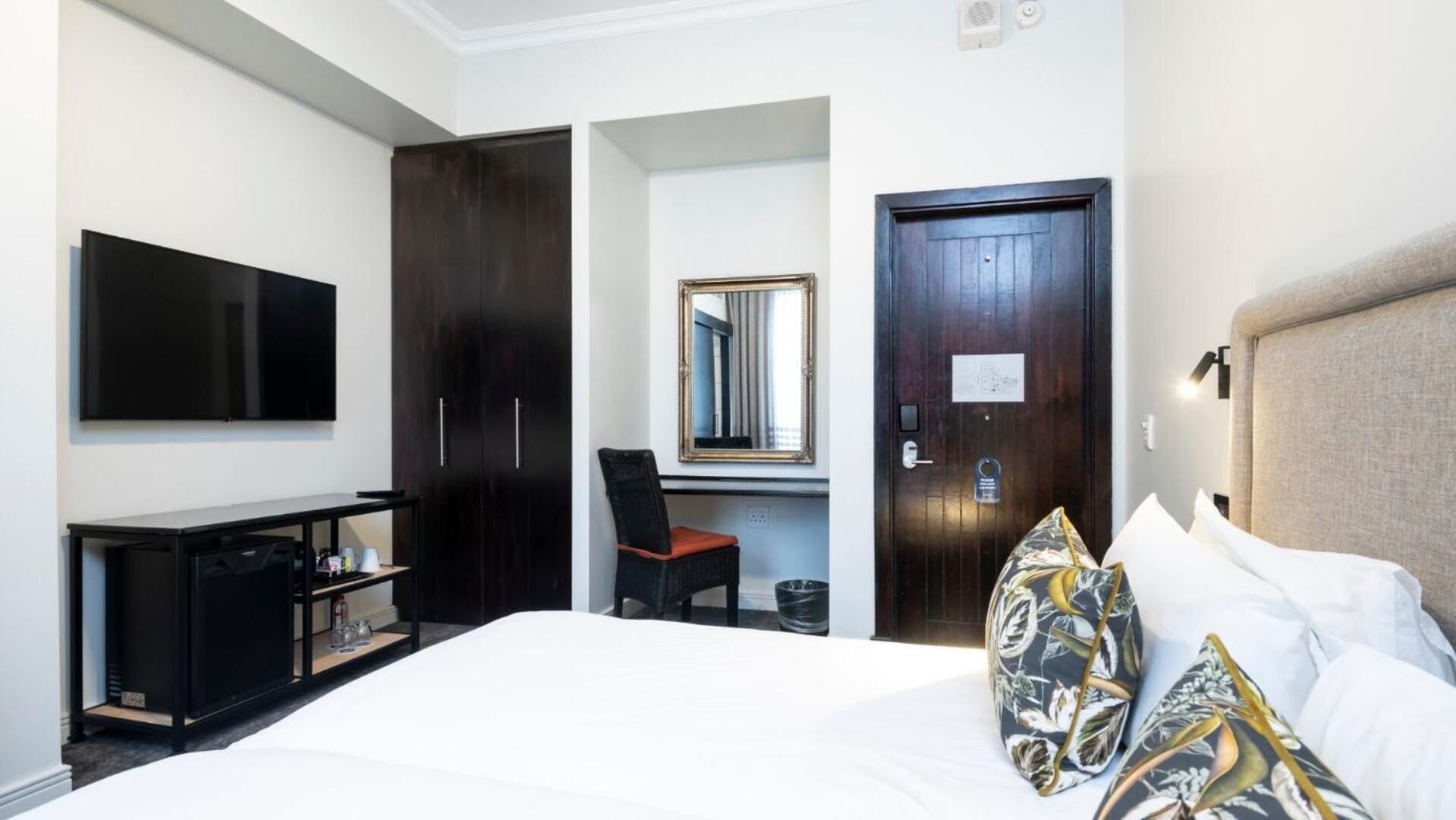 A hotel room with a large bed, a TV mounted on the wall, a desk with a chair, a wardrobe, and a door with a mirror.